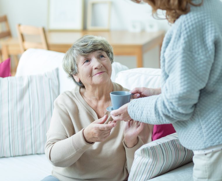 Betreuungskraft reicht einer Seniorin eine Tasse Kaffee im Wohnzimmer